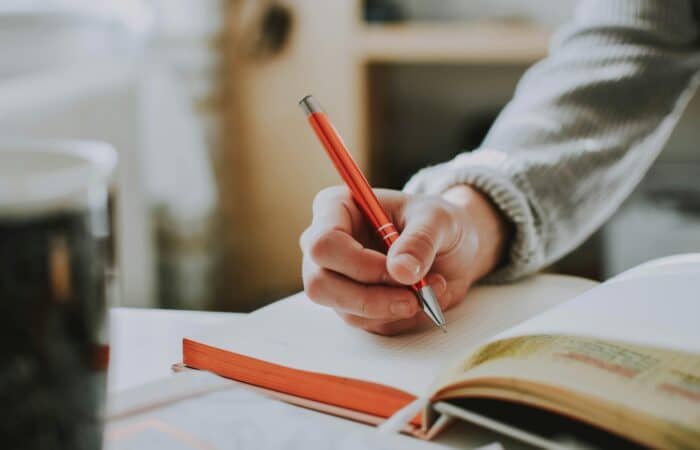 A person's hand holding an orange pen on a white page in a notebook, getting to write.