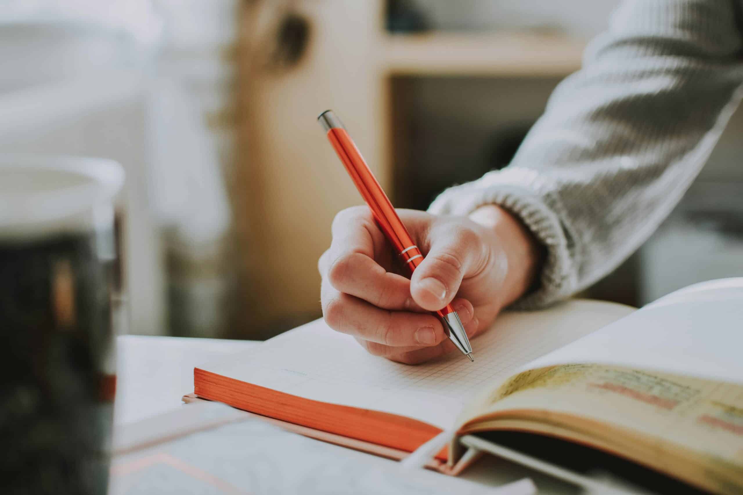 A person's hand holding an orange pen on a white page in a notebook, getting to write.