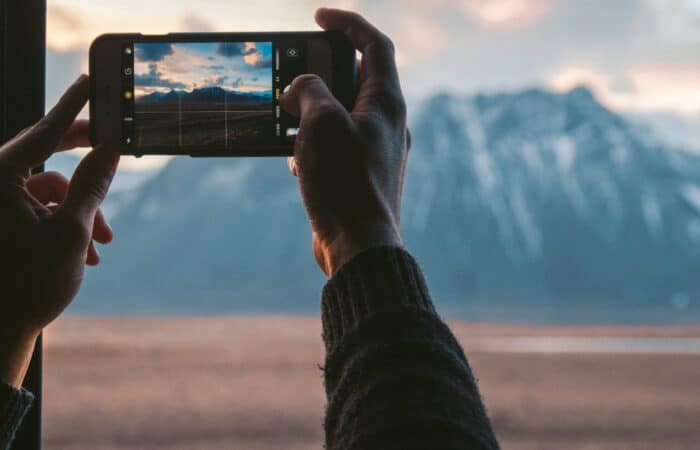 A man takes a photo of a mountain from his window. The sun is setting and there is snow on the peaks.