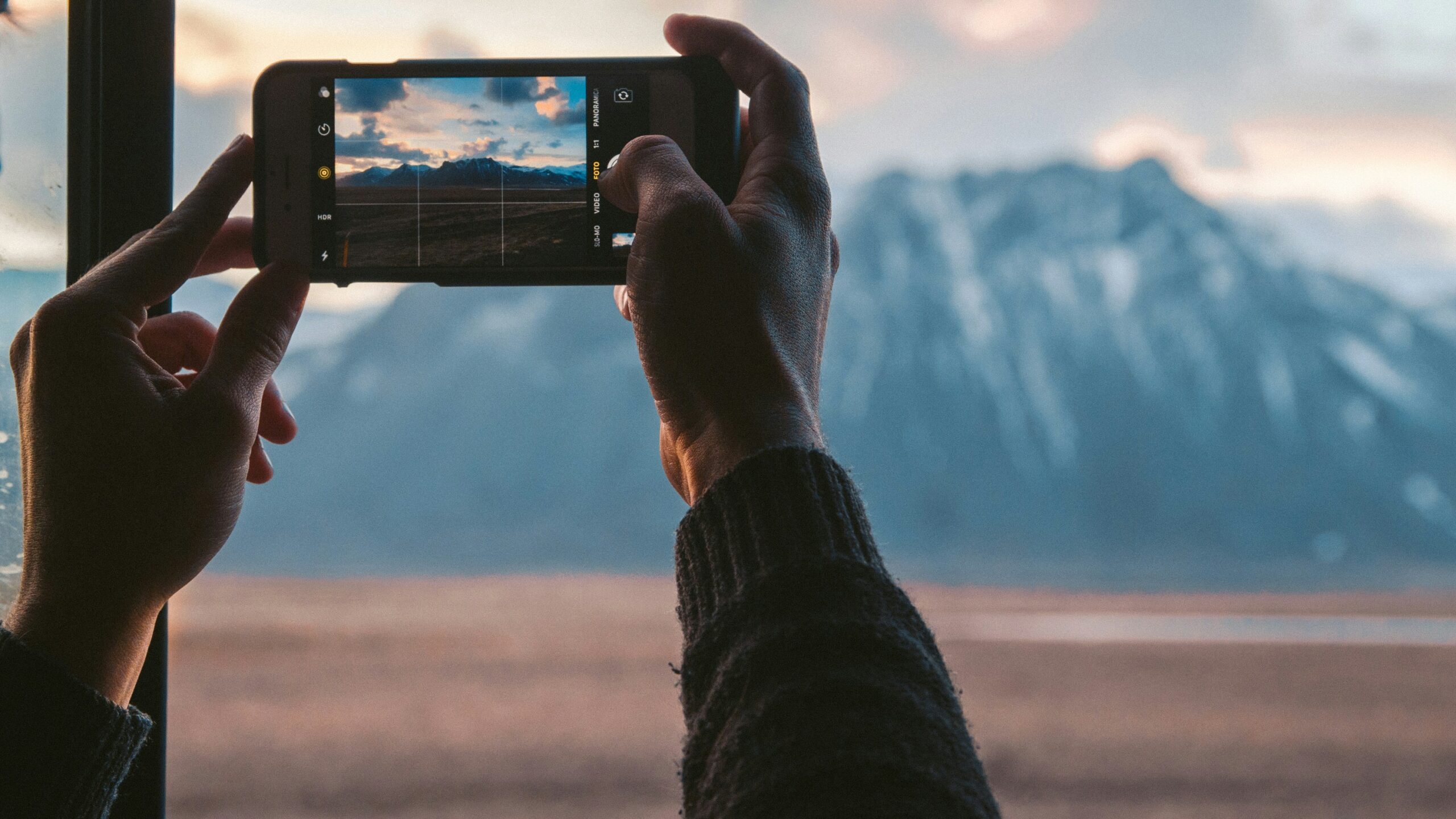 A man takes a photo of a mountain from his window. The sun is setting and there is snow on the peaks.