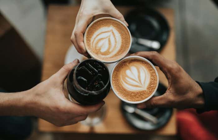 Three people clinking coffee cups, two lattes and one iced coffee, in a circle above a wooden table.