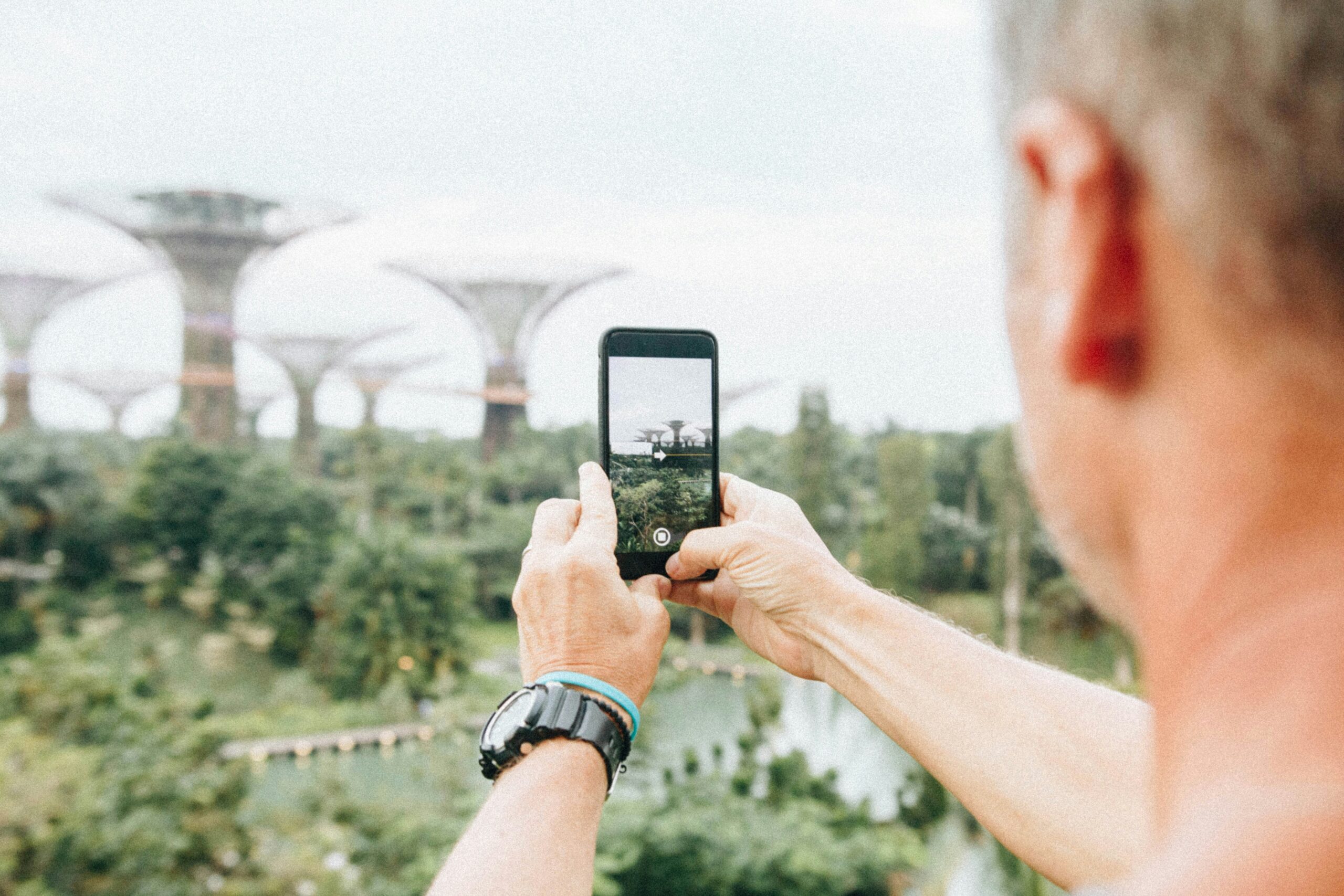 Man takes a picture of futuristic buildings with his mobile phone while standing in greenery.