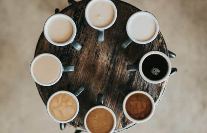 8 coffee cups in a circle on a small table. Each cup showing a different shade of coffee, some very milky, but all different strengths.
