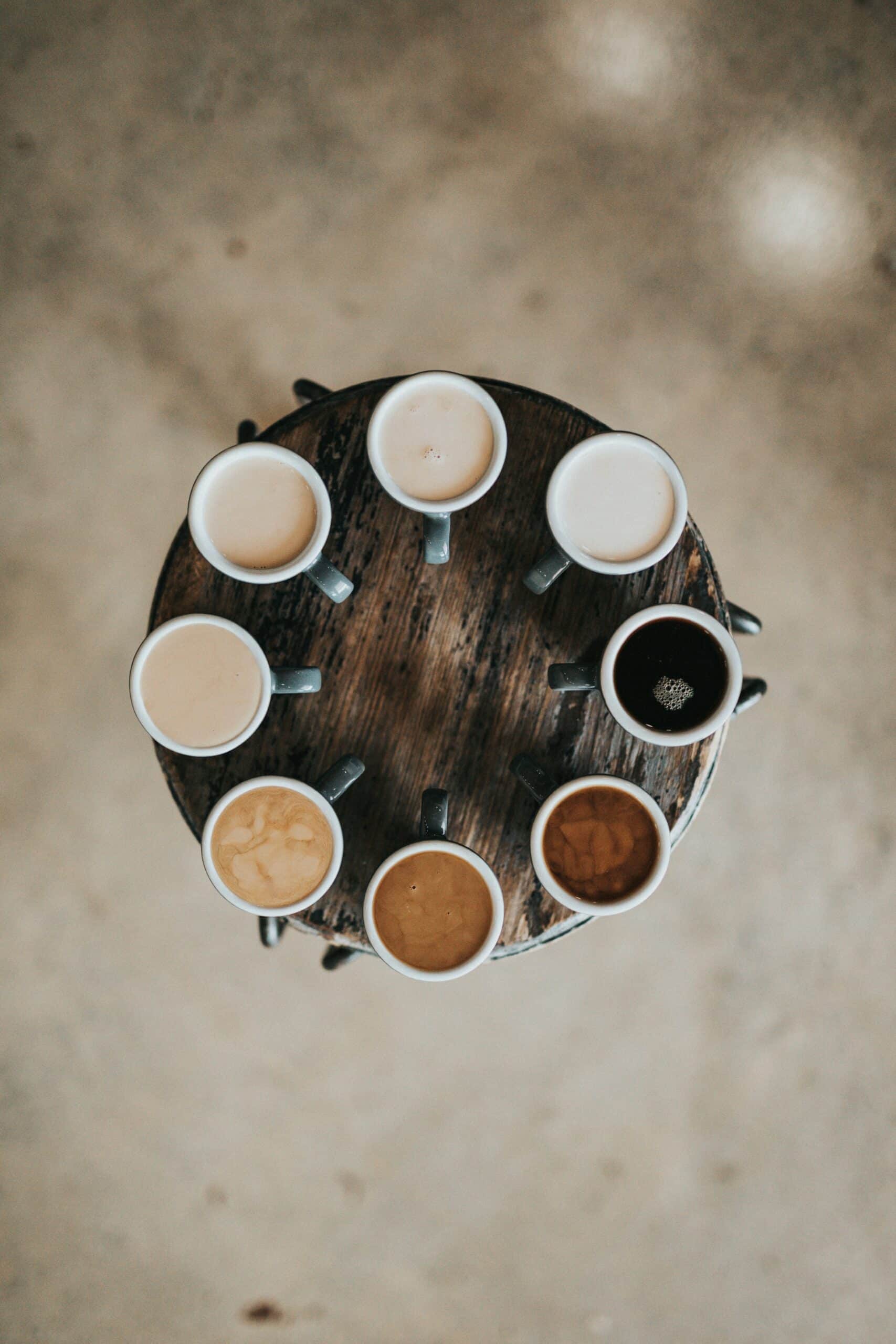 8 coffee cups in a circle on a small table. Each cup showing a different shade of coffee, some very milky, but all different strengths.