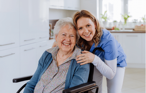 A young woman carer and an older woman caree who is sitting in wheelchair in a clean home. Both of them are smiling at the camera.